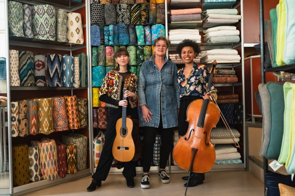 Three women with stand in front of colorful textiles, one person has an acoustic guitar and another a cello.