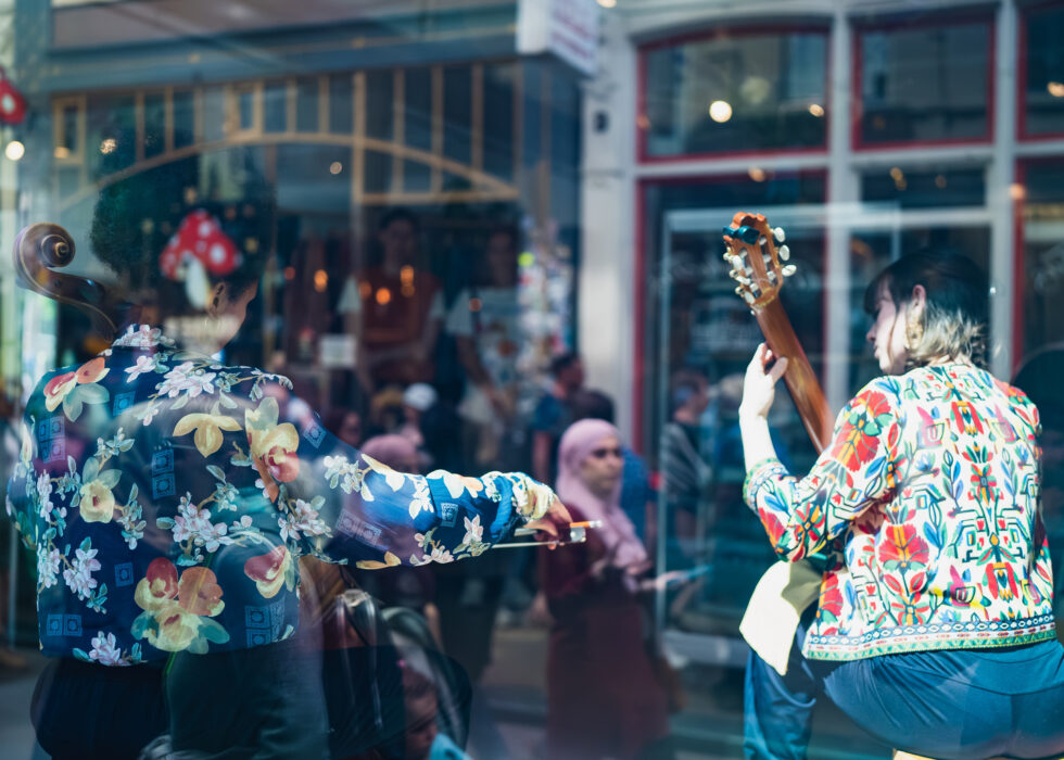 Two musicians play cello and guitar behind a window, floral shirts.