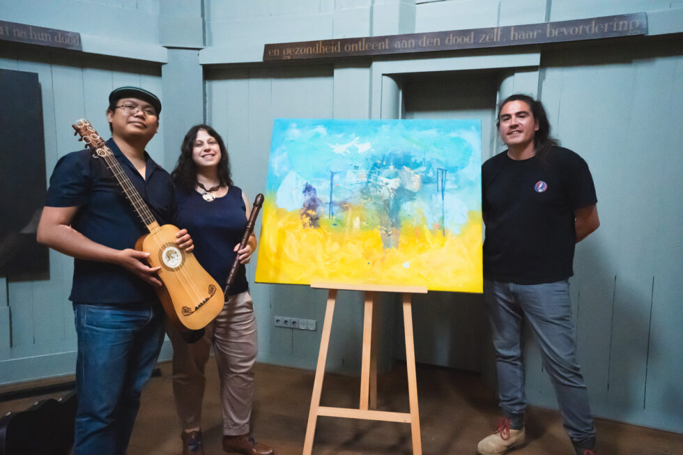 3 people pose with art work; one musician with a baroque guitar and one with a flute.