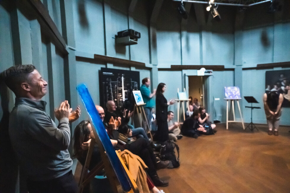 Audience applauds at a performance event, paintings on display.