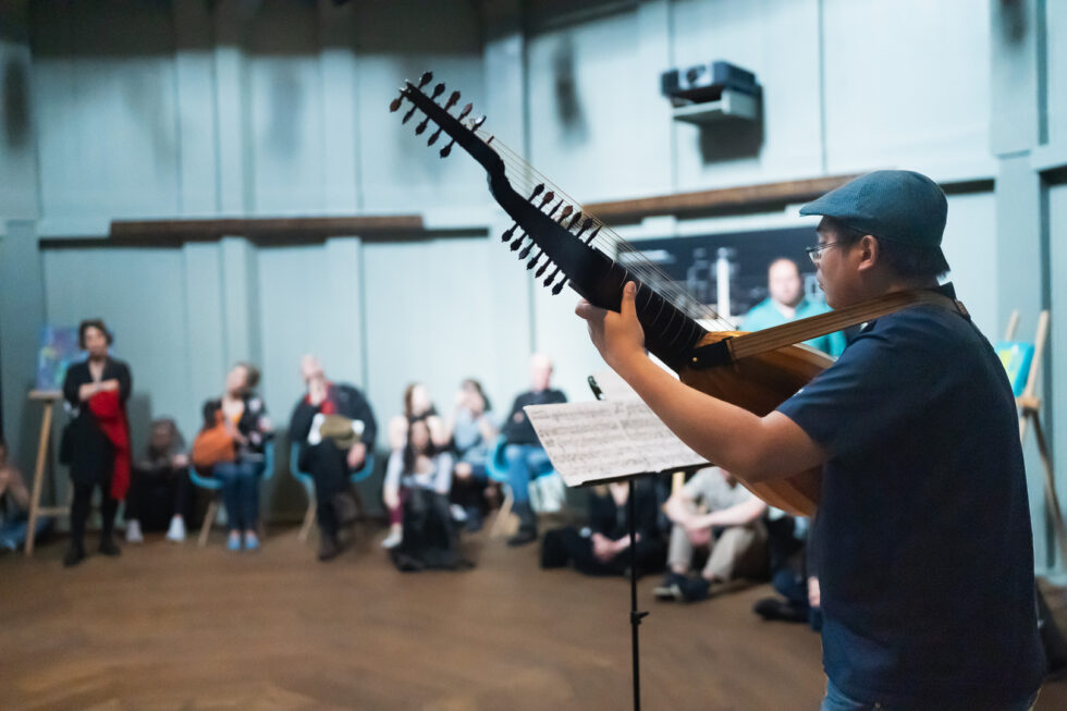Musician plays lute for audience, surrounded by audience and art