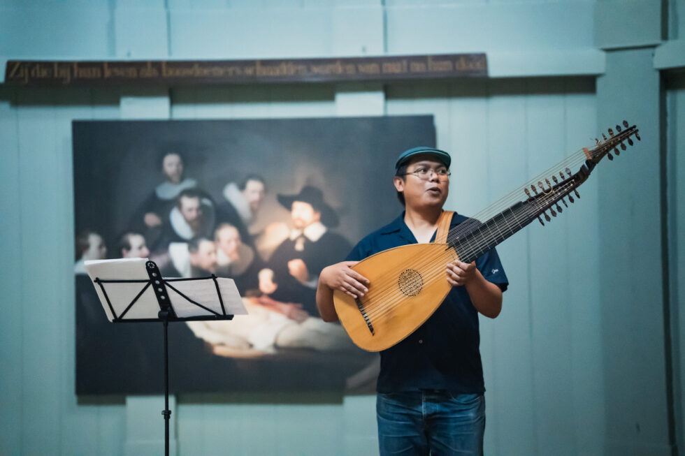 Musician playing a theorbo lute in front of a Rembrandt painting.
