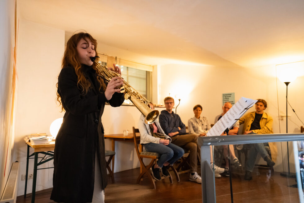 Woman plays saxophone for small audience in warm, inviting room.
