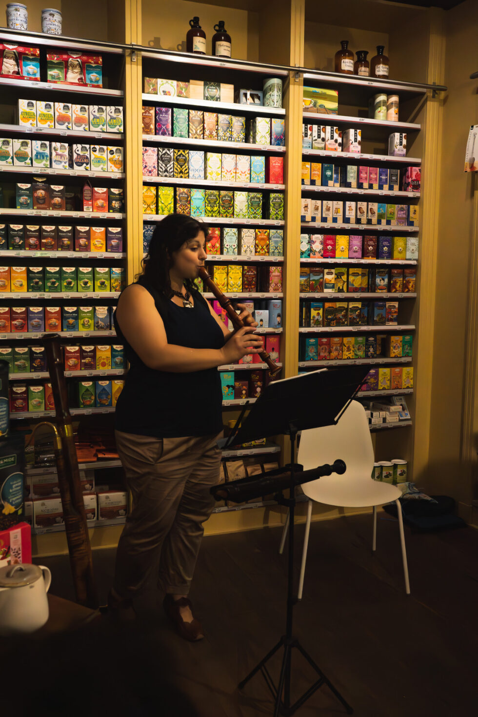 Woman plays recorder in tea shop with shelves of colorful tea boxes.