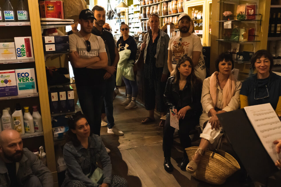 Group of people gathered in a shop, some sitting, some standing, watching a performance.