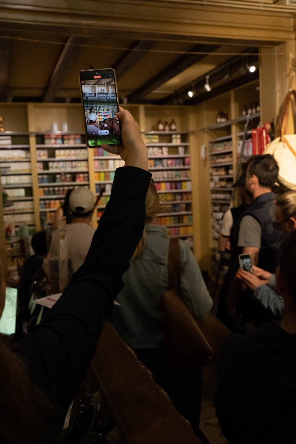 Person video recording with their phone, a performance that is happening in a small store, with audience members looking on.