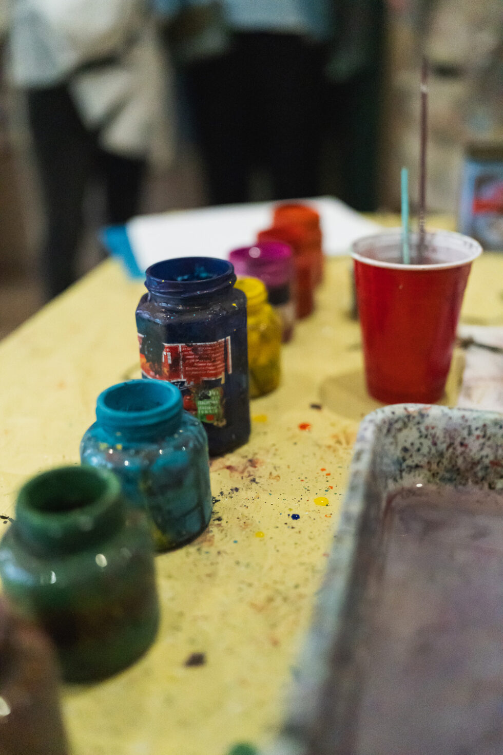 Colorful paint jars and a red cup on a splattered yellow table.