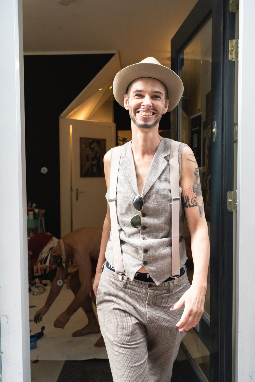 Smiling person in vest and hat stands in doorway with art studio interior