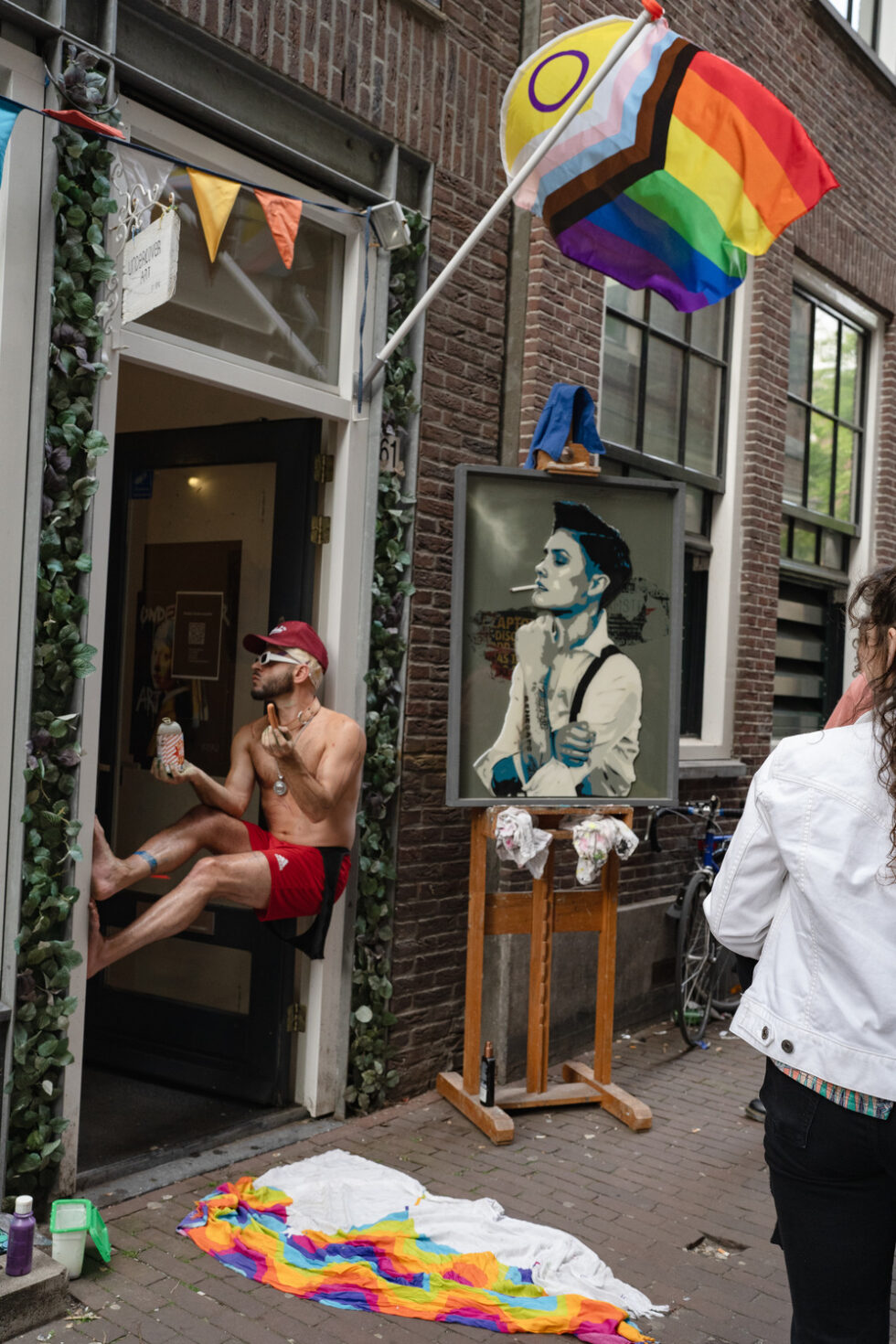 Man in doorway under Pride flag, Amsterdam. David Bowie art displayed. Street scene.