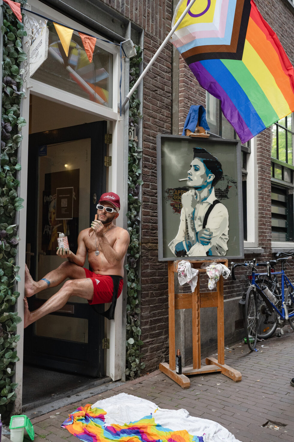 Shirtless performance artist propped up in doorway with pride flags and David Bowie pop-art. Amsterdam.