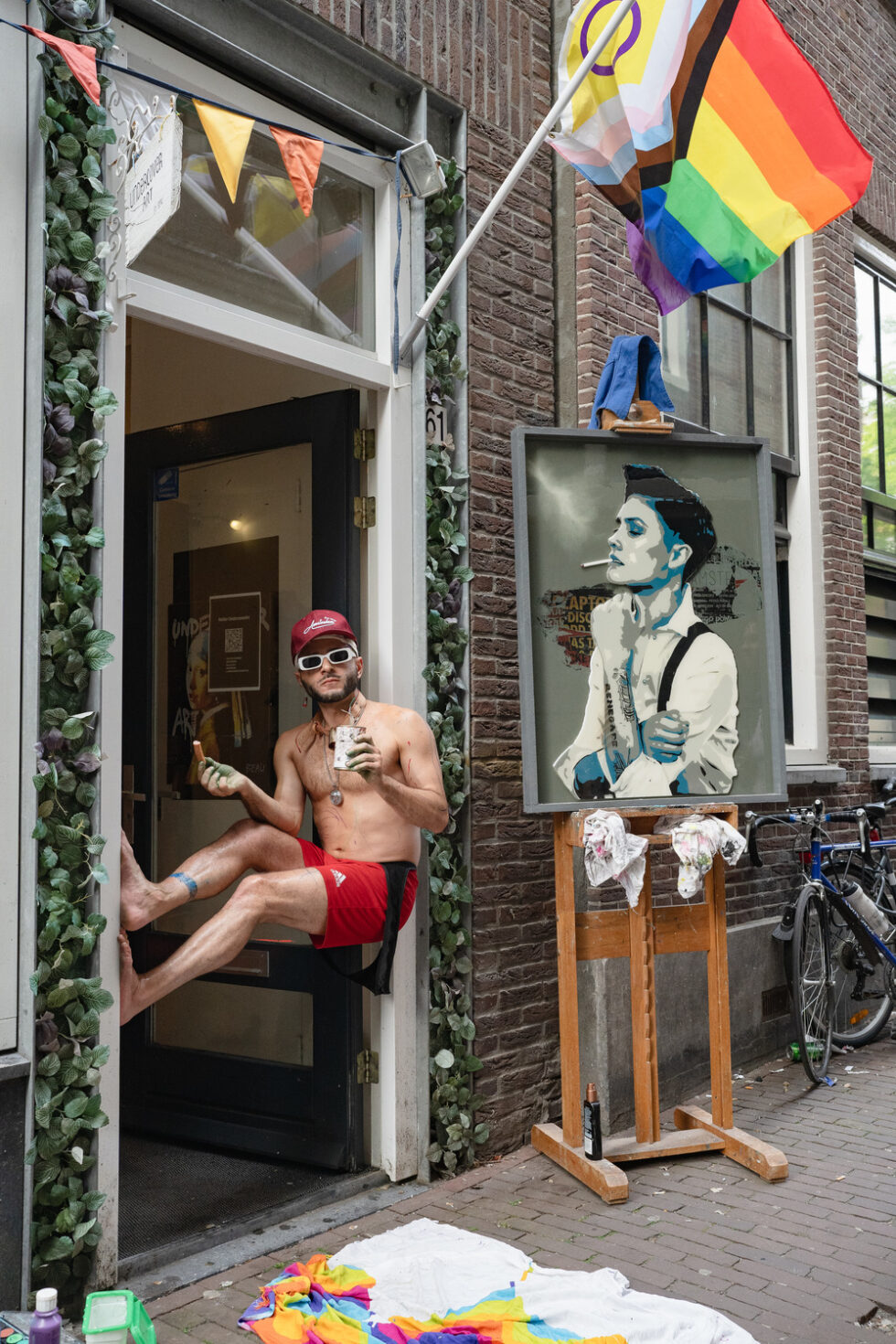 Shirtless performance artist propped up in doorway with pride flags and David Bowie pop-art.