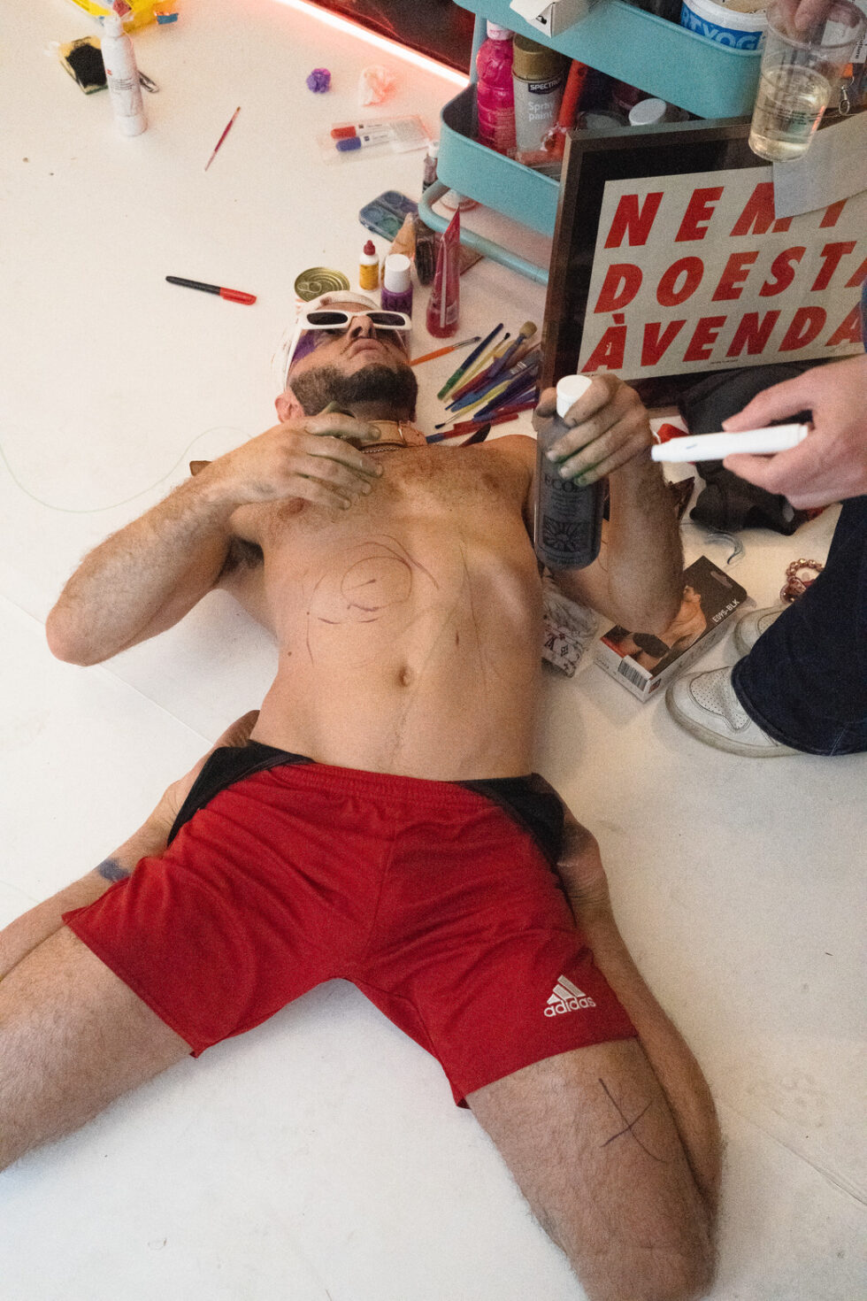Man in red shorts lying on floor, body art being applied.