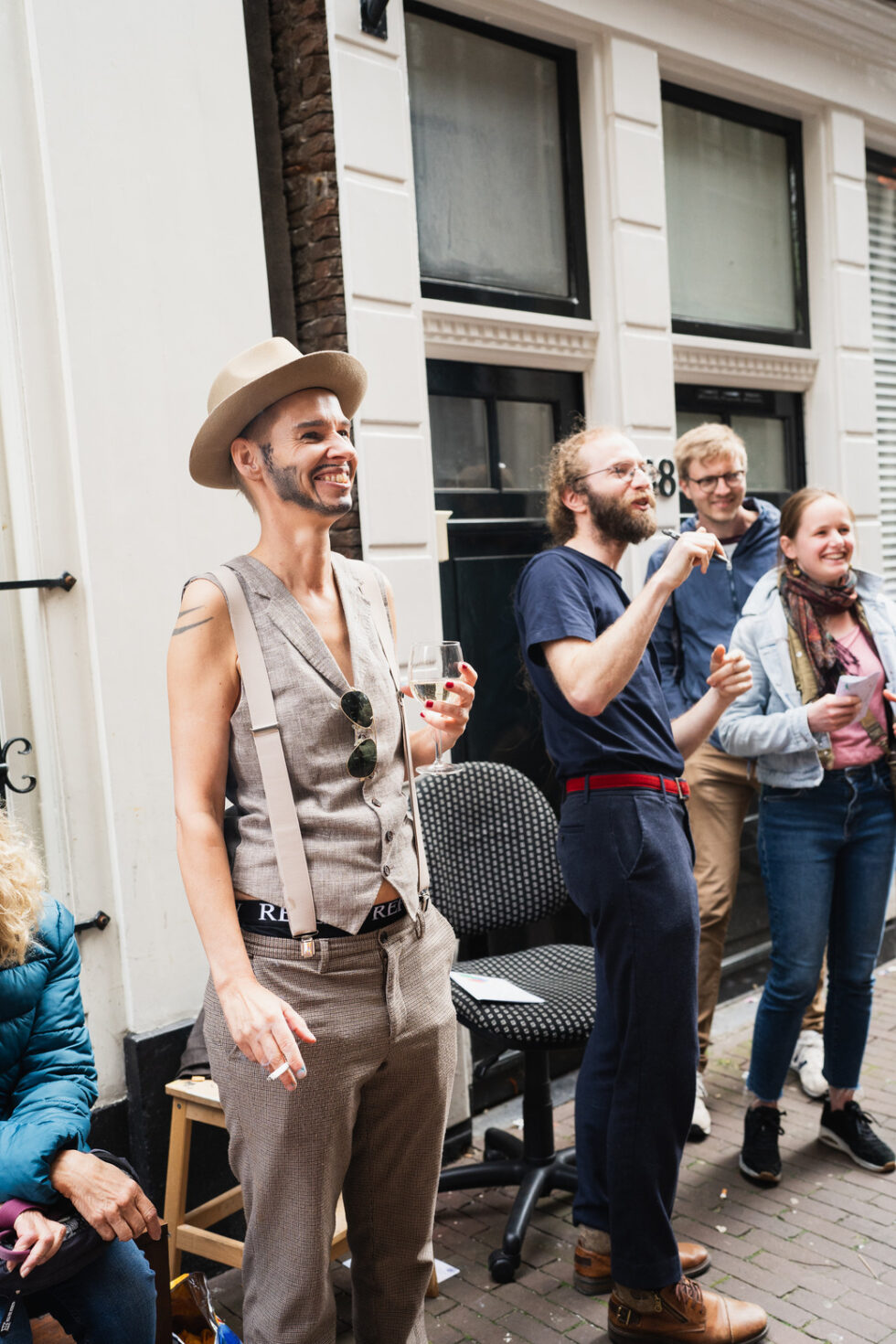 Person in hat and vest with wine glass, smiling at outdoor event with people and building in background.