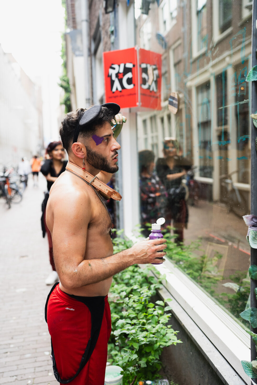 Shirtless man performance artist, making drawings on a window with the Red Light Arts and Culture sign in the background.