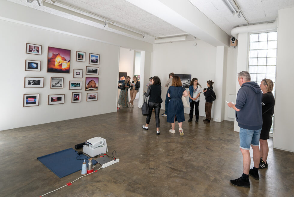 Gallery visitors viewing framed art on white walls, with a projector and mat in the foreground.