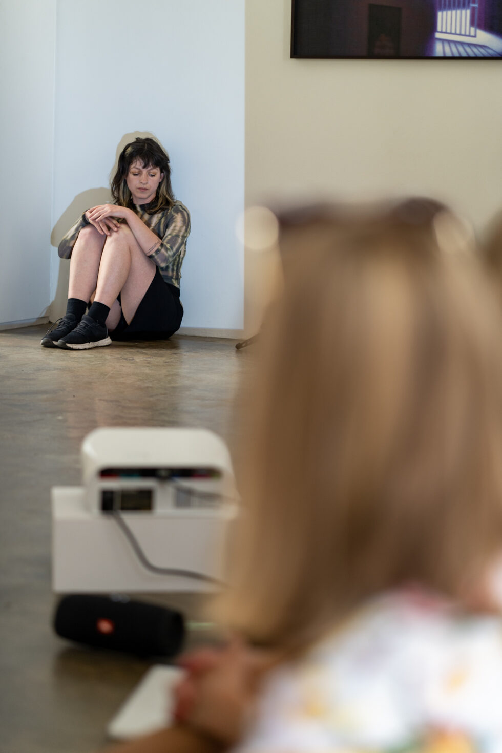 Woman sitting in corner, looking down. Art installation with projector in foreground.