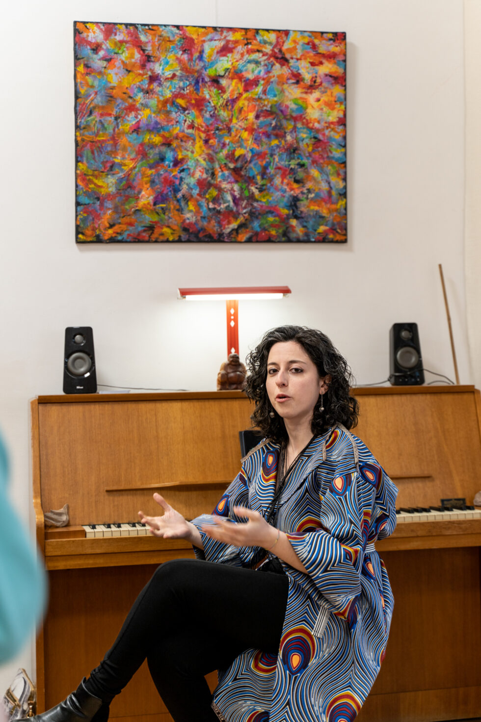 Woman in patterned coat sits by piano with colorful abstract art above.