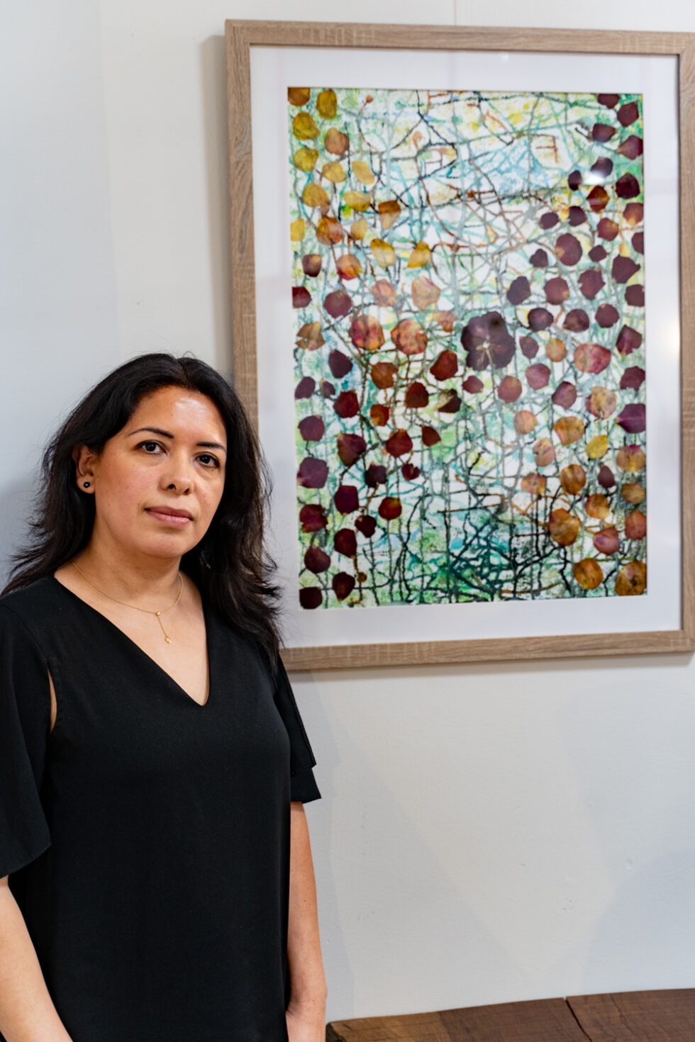 An artists stands beside their framed abstract art with organic shapes in browns, reds, yellows, and blues.