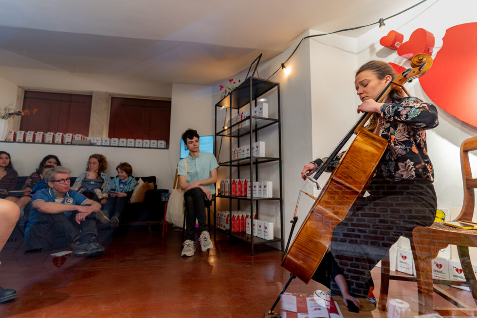 Cellist performing for a small audience in an intimate setting, with shelves of products in the background.