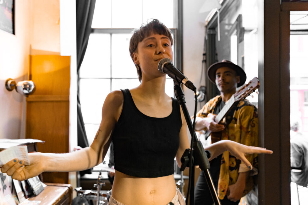 Announcer talking into microphone with guitarist behind, live music event