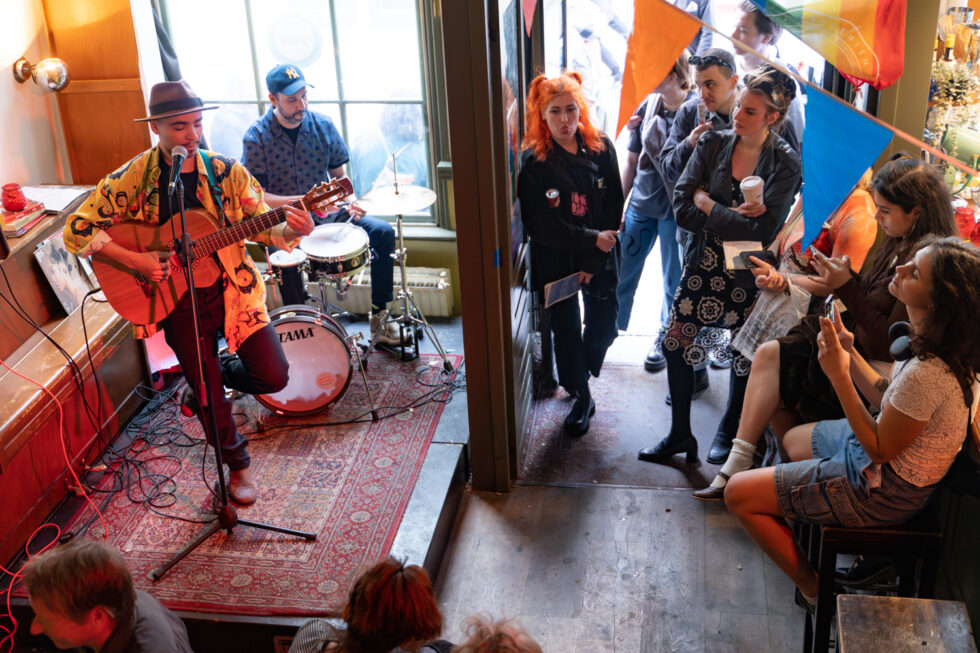 Live music performance: Musician playing guitar, drummer behind. Audience sitting at bar watching on