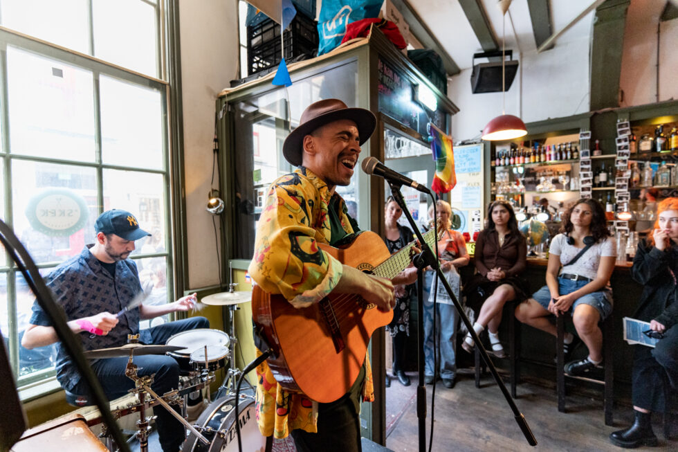 Live music performance: Musician playing guitar, drummer behind. Audience sitting at bar watching on