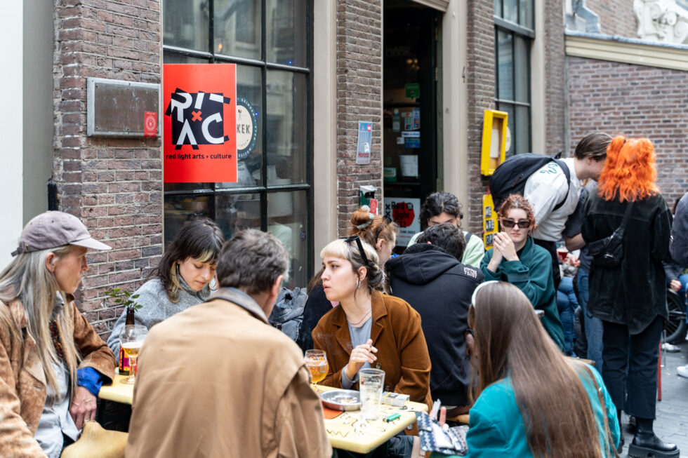 Group of people sitting at tables outside a cafe with red sign reading RLAC